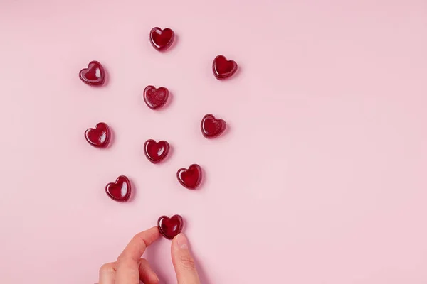 Hand hält eine der verstreuten roten Gelee-Bonbons in Herzform auf rosa Hintergrund — Stockfoto
