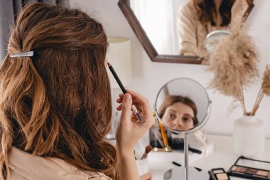 Genç bir kadın yüzüne makyaj yapıyor, yuvarlak aynaya bakıyor.