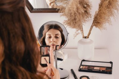 Genç bir kadın yüzüne makyaj yapıyor, yuvarlak aynaya bakıyor.