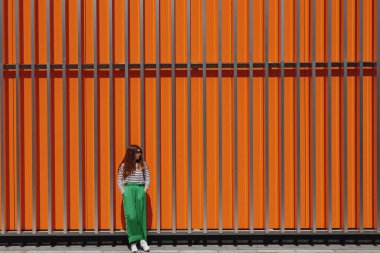 Metall turuncu duvarın yanında kot pantolon giymiş şık bir kadın portresi.