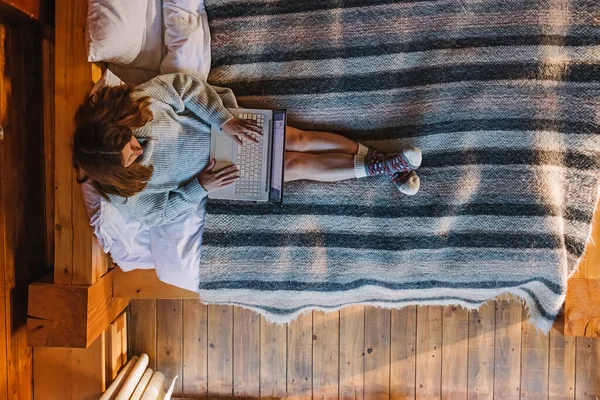 Vrouw in trui en warme sokken werkend op laptop zittend op gezellig bed — Stockfoto