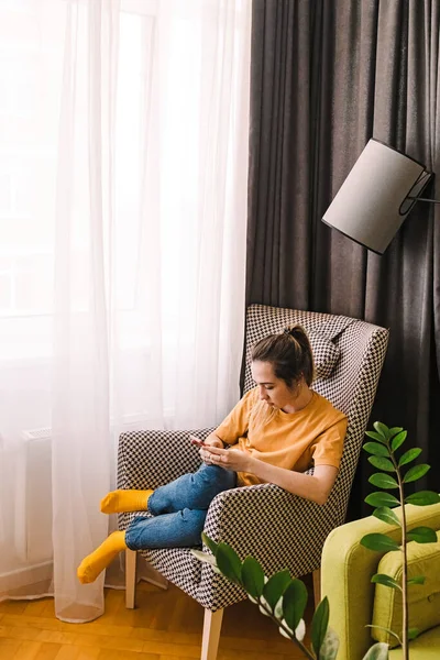 Giovane donna con smartphone seduta nella comoda poltrona di casa — Foto Stock