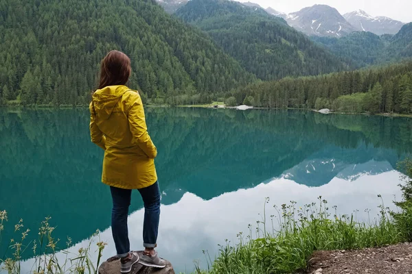 穏やかな山の湖のほとりに立つ黄色のレインコートの女性旅行者 — ストック写真