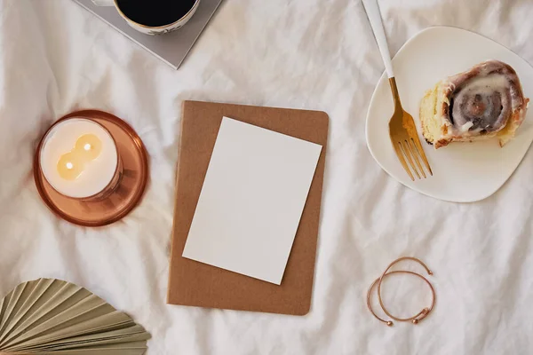 Tarjeta de papel maqueta en la cama con café, vela y rollo de canela —  Fotos de Stock