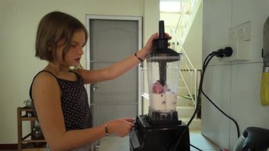 Genç bir kızın modern mutfağındaki güçlü bir blender 'da sağlıklı meyve suyu hazırlarken parlak ve enerjik bir pozu. Malzemeleri harmanlarken aktif olarak gösteriliyor.