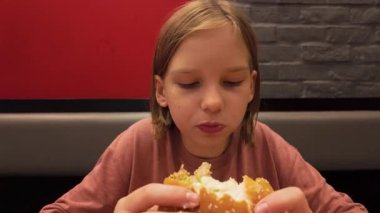 Neşeli, samimi bir fotoğraf, genç bir kızı, büyük, cezbedici bir hamburgerden tatmin edici bir ısırık alırken yakalar. Belli ki sıradan bir ortamda yemeğinin tadını çıkarır. Görüntülerde gençlik kültürü temaları vurgulanıyor