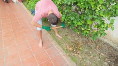 Sıradan pembe tişörtlü, kot şortlu ve yeşil eldivenli bahçedeki yaprakları tırmıklayan genç sakallı adam. Açık havaya odaklanmış, yeşil çalıların ve ağaçların yakınındaki fayanslı bir patikada yalınayak duruyor.