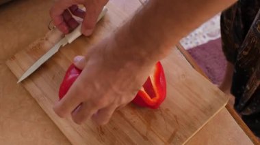 Canlı ve detaylı bir yakın çekim, kısa sakallı 38 yaşındaki Avrupalı bir adamın eline odaklanıyor. Tahta kesme tahtasındaki parlak kırmızı biberi dikkatle dilimliyor ve taze salata hazırlıyor.