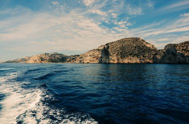 Akdeniz kıyı şeridi boyunca uzanan kireçtaşı kayalıkları derin mavi dalgalar ve bulutlu gökyüzü ile denizden görülüyor.