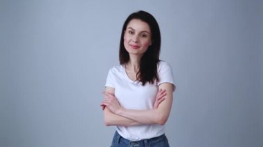 Mutlu genç esmer kadın stüdyoda kollarını göğsünde kavuşturuyor. Beyaz tişörtle poz veren güzel beyaz kadın modelinin portre görüntüleri, mutlu, dişlek gülüşlü kot pantolon. 30 'lu yaşlarda çekici beyaz bir kız.