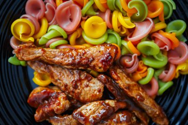 Karamel soslu kızarmış domuz eti ve siyah seramik tabakta servis edilen çok renkli makarna. Yukardan düz bir şekilde çekilmiş. Ev mutfağında akşam yemeği için nefis bir bbq yemeği.