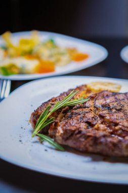 Restoranda akşam yemeği için ızgarada pişmiş nefis pirzola bifteği. Tavada ızgara yapılmış ve biberiyeli beyaz tabakta servis edilen gurme bifteğinin dikey yakın çekimi. Lezzetli kırmızı et filetosunun tadını çıkarın.