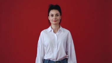 Kamerada gülen sevimli, genç bir kadın. Beyaz gömlek ve kot pantolon giyen beyaz bir kadın stüdyoda kırmızı arka planda beyaz dişlerle gülüyor ve gülüyor. 30 'ların ortasında dost canlısı bir model. 