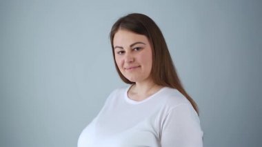 Stüdyoda arkadaş canlısı bir gülümsemeyle poz veren doğal görünüşlü, iri yarı, uzun saçlı, klasik beyaz tişörtlü, arkadaş canlısı bir genç kadın. 