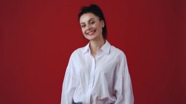 Kamerada gülen sevimli, genç bir kadın. Beyaz gömlek ve kot pantolon giyen beyaz bir kadın stüdyoda kırmızı arka planda beyaz dişlerle gülüyor ve gülüyor. 30 'ların ortasında dost canlısı bir model. 