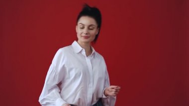 Kamerada gülen sevimli, genç bir kadın. Beyaz gömlek ve kot pantolon giyen beyaz bir kadın stüdyoda kırmızı arka planda beyaz dişlerle gülüyor ve gülüyor. 30 'lu yaşlarda dost canlısı bir model 