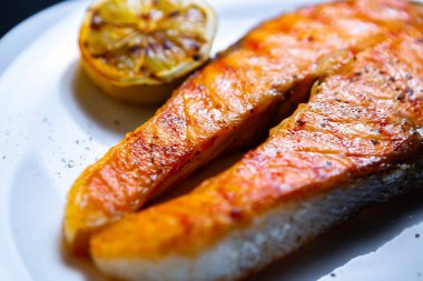 Akşam yemeği için ızgarada pişmiş somon balığı bifteği. Deniz ürünleri restoranında ızgara yapılan ve limonlu beyaz seramik tabakta servis edilen lezzetli kırmızı balık filetosu. Kraliyetin ücretsiz küratörlü yemek resimlerini indirin.