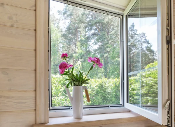 Ventana blanca con mosquitero en una casa de madera rústica con vistas al jardín. Ramo de peonías rosadas en una regadera en el alféizar de la ventana Fotos De Stock