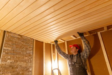 İşçi, başındaki ahşap tavan panelini yalıtılmış odanın içine yerleştiriyor. Kereste çerçevesi görünür, iç yenileme süreci, el işçiliği, işçilik ve konut inşaatı çalışmaları sürüyor.