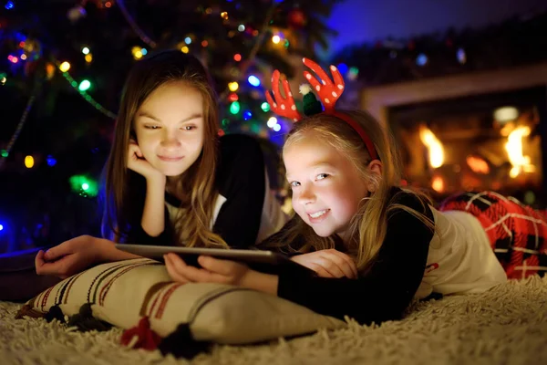 平安夜 两个可爱的妹妹在温暖舒适的客厅里用平板电脑在家里壁炉边 冬夜与家人和孩子呆在家里 — 图库照片