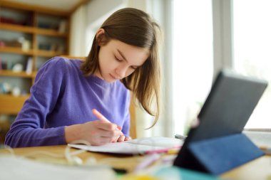 Liseli kız evde dijital tabletle ödevini yapıyor. Çocuk çalışmak için aletler kullanıyor. Çocuklar için eğitim ve uzak mesafe eğitimi. Karantina sırasında evde eğitim. Evde kalıp eğlenin..