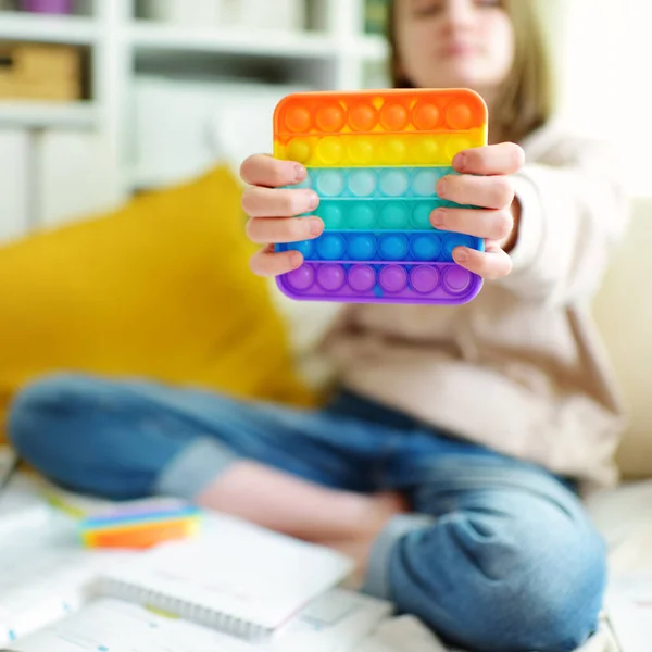 自宅で勉強しながら虹色のポップ イットのフィデット トイで遊ぶ10代の女の子 トレンディなストレスと不安救済フィギングゲームを持つ10代の子供 感覚シリコーンおもちゃのディンプルをポップ — ストック写真