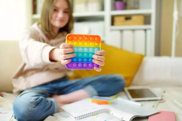 Teenage girl playing with rainbow pop-it fidget toy while studying at home. Teen kid with trendy stress and anxiety relief fidgeting game. Popping the dimples of sensory silicone toy.