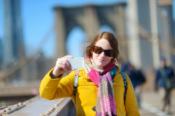 Beautiful young woman taking selfie with smartphone on Brooklyn Bridge, New York, at sunny spring day