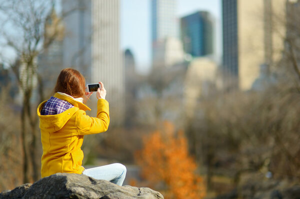 Beautiful young woman taking a photo of skyscrapers while sitting on a rock in Central Park, New York