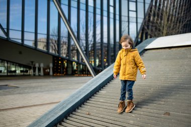 Hardal ceketli ve kot pantolonlu çocuk Vilnius 'taki bir şehir parkında modern bir ahşap binaya tırmanıyor. Kentsel kamusal alanda neşeli bir hareket ve merak anı.