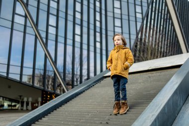 Hardal ceketli ve kot pantolonlu çocuk Vilnius 'taki bir şehir parkında modern bir ahşap binaya tırmanıyor. Kentsel kamusal alanda neşeli bir hareket ve merak anı.