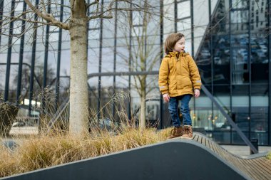 Hardal ceketli ve kot pantolonlu çocuk Vilnius 'taki bir şehir parkında modern bir ahşap binaya tırmanıyor. Kentsel kamusal alanda neşeli bir hareket ve merak anı.