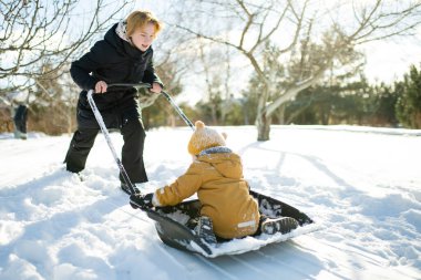 Hardal ceketli, sarı şapkalı, kar küreğinde gezen, karda alçakta oturan gülen çocuk. Doğaçlama kış kızağı, parlak soğuk güneş ışığında neşeli bir doğallık..