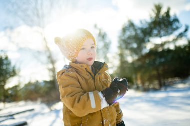 Hardal ceketli gülümseyen çocuk ve örgü şapkalı çocuk taze karda çömeliyor, soğuktan yanakları pembeleşiyor. Kış eğlencesi, açık hava ışığında basit çocukluk özgürlüğü..