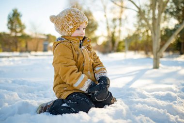 Hardal ceketli gülümseyen çocuk ve örgü şapkalı çocuk taze karda çömeliyor, soğuktan yanakları pembeleşiyor. Kış eğlencesi, açık hava ışığında basit çocukluk özgürlüğü..