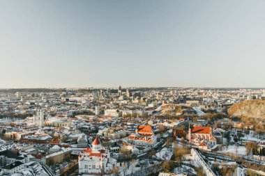 Kış güneşinde parlayan kiliseleri ve kırmızı çatıları olan karla kaplı Vilnius Eski Kasabası 'nın insansız hava aracı görüntüsü. Tarihsel şehir manzarası, sakin mevsimsel uyum içinde modern ufuk çizgisiyle buluşur..