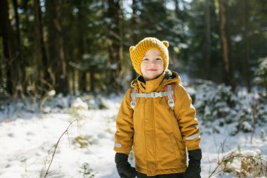 Hardal ceketli ve sarı örgü şapkalı bir çocuk sırt çantalı, güneş ışığına gülümseyen bir çocuk. Kış macera havası, vahşi doğadaki genç kaşif enerjisi..
