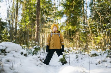 Hardal ceketli ve sarı örgü şapkalı bir çocuk sırt çantalı, güneş ışığına gülümseyen bir çocuk. Kış macera havası, vahşi doğadaki genç kaşif enerjisi..