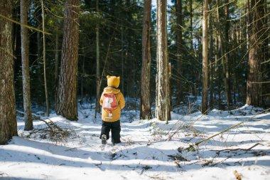 Hardal ceketli ve sarı örgü şapkalı bir çocuk sırt çantalı, güneş ışığına gülümseyen bir çocuk. Kış macera havası, vahşi doğadaki genç kaşif enerjisi..