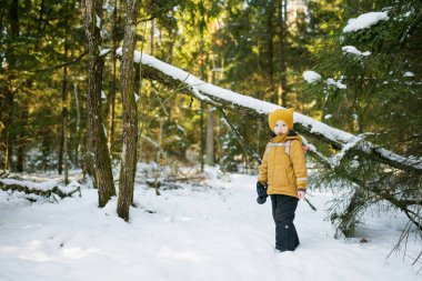 Hardal ceketli ve sarı örgü şapkalı bir çocuk sırt çantalı, güneş ışığına gülümseyen bir çocuk. Kış macera havası, vahşi doğadaki genç kaşif enerjisi..