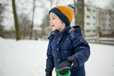 Donanma ceketli ve örgü şapkalı bir çocuk karlı bir şehir parkında duruyor, yüzü kar taneleriyle kaplanmış. Sakin bir kış anı, sakin bir varlık yumuşak şehir karları arasında.