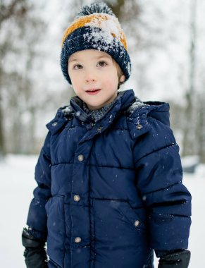 Child in a navy jacket and knit hat stands in a snowy city park, face dusted with snowflakes. A calm winter moment, quiet presence amid soft urban snowfall.