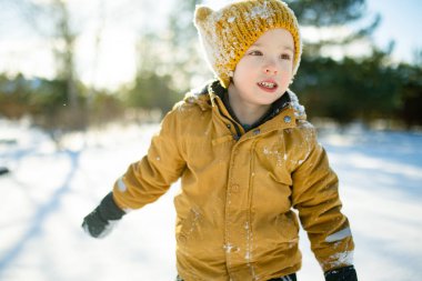 Hardal ceketli gülümseyen çocuk ve örgü şapkalı çocuk taze karda çömeliyor, soğuktan yanakları pembeleşiyor. Kış eğlencesi, açık hava ışığında basit çocukluk özgürlüğü..