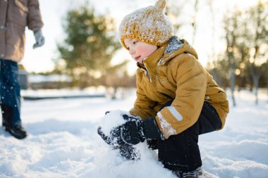 Hardal ceketli gülümseyen çocuk ve örgü şapkalı çocuk taze karda çömeliyor, soğuktan yanakları pembeleşiyor. Kış eğlencesi, açık hava ışığında basit çocukluk özgürlüğü..