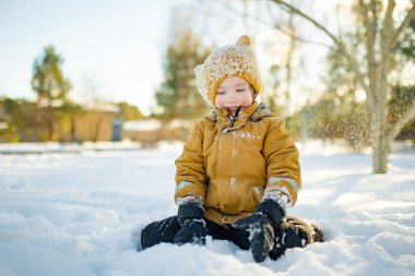 Hardal ceketli gülümseyen çocuk ve örgü şapkalı çocuk taze karda çömeliyor, soğuktan yanakları pembeleşiyor. Kış eğlencesi, açık hava ışığında basit çocukluk özgürlüğü..