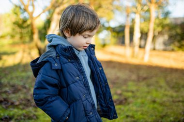 Mavi kapüşonlu ve kabarık ceketli gülümseyen çocuk gri bir günde yeşil bir bahçede duruyor. Hafif Avrupa kışı ya da sonbahar sonu, mevsimler arasında sakin bir an.