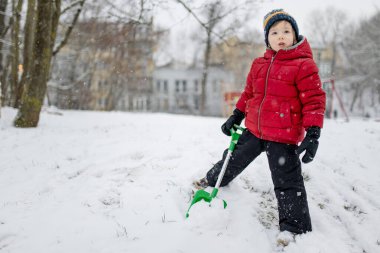 Düşen karda kırmızı ceketli bir çocuk yeşil bir oyuncak küreği yere bastırıyor. Karlı bir şehir gününde sakin bir kararlılıkla bir kış oyunu..