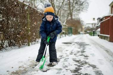 Kışın giyinmiş bir çocuk, yeşil bir kar küreğiyle karlı bir şehir kaldırımında çitin yanında duruyor. Sıradan bir kış günü, hareketli ve sessiz bir mevcudiyetle.
