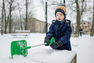 Kışın giyinmiş bir çocuk, yeşil bir kar küreğiyle karlı bir şehir kaldırımında çitin yanında duruyor. Sıradan bir kış günü, hareketli ve sessiz bir mevcudiyetle.