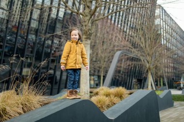 Hardal ceketli ve kot pantolonlu çocuk Vilnius 'taki bir şehir parkında modern bir ahşap binaya tırmanıyor. Kentsel kamusal alanda neşeli bir hareket ve merak anı.